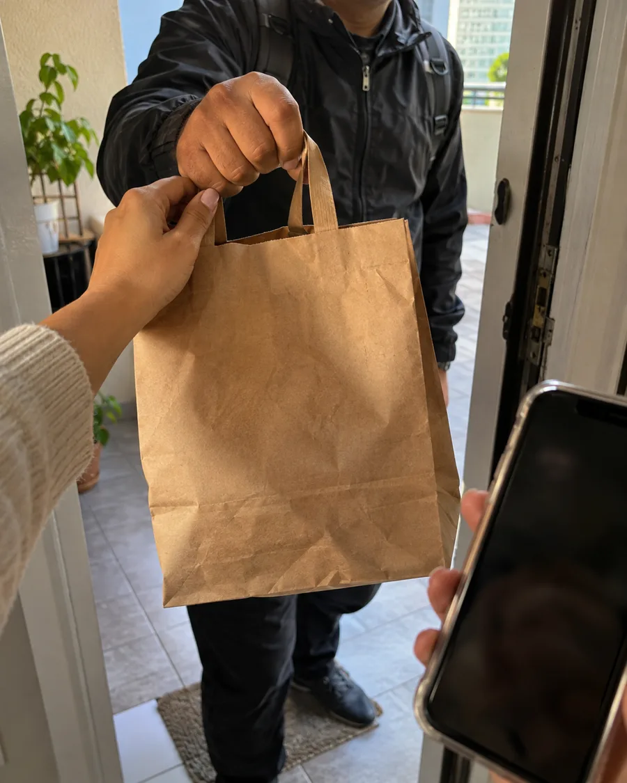 Entregador passa uma sacola de papel pela porta de um apartamento enquanto a pessoa segura o celular